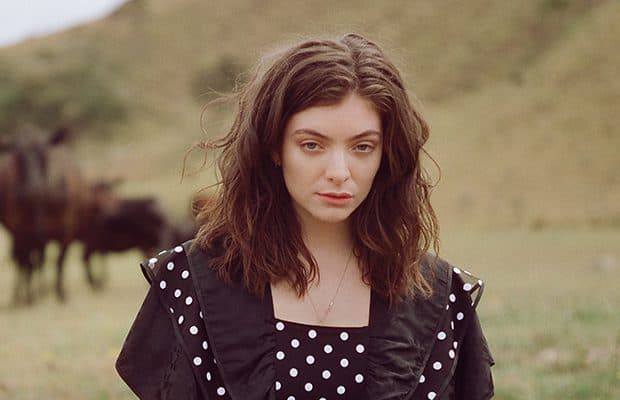 A picture of Lorde looking into the camera. In the background is greenery and grass.
